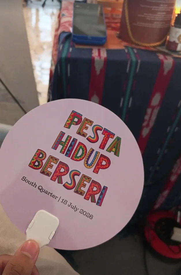 A hand holding a colorful Pesta Hidup Berseri fan dated 18 July 2026 at South Quarter